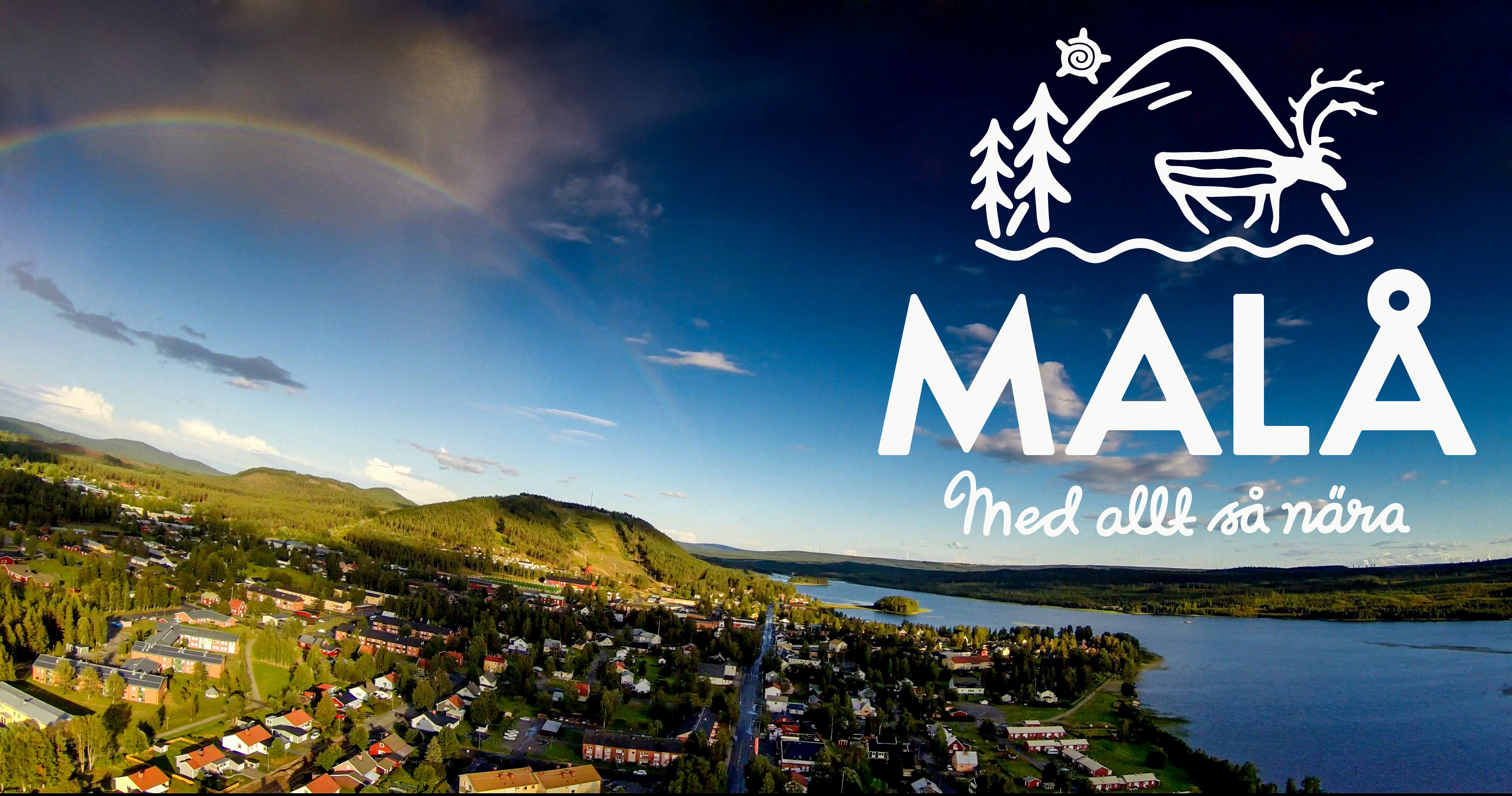 Flygbild över Malå samhälle. Regnbåge över berg och skog i fjärran. Malåns vatten. 